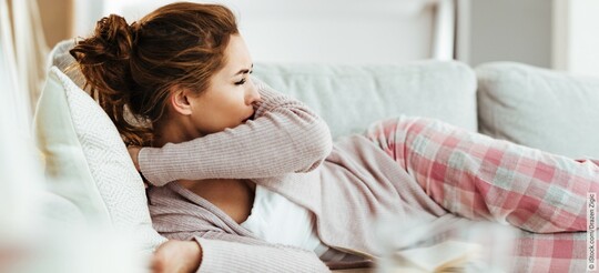 Verschleppte Erkältung - Frau liegt im Schlafanzug auf der Couch und hustet in ihren Ellenbogen