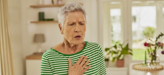 Frau mit Asthma bronchiale steht im Wohnzimmer und legt die Hand auf den Brustkorb. An ihrem Gesichtsausdruck ist zu erkennen, dass sie Probleme beim Atmen hat.