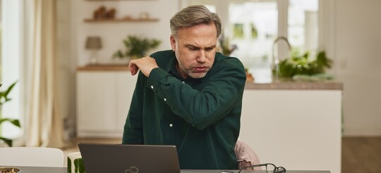 Raucherhusten - Mann sitzt im Büro vor seinem Laptop und hustet in seinen Ellenbogen. Sein Gesicht sieht schmerzverzerrt aus