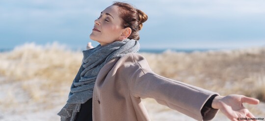 Frau mit brünetten Haaren steht im Winter am Strand in den Dünen und mit ausgebreiteten Armen. Sie macht eine Klimatherapie und genießt sichtlich das Reizklima der frischen und salzhaltigen Meeresluft.