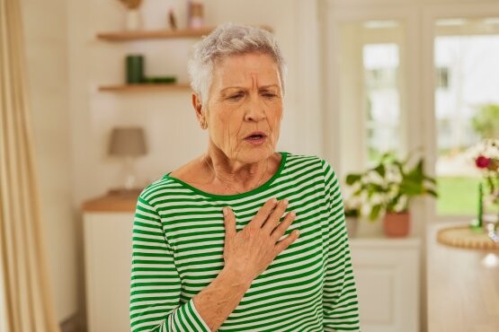 Ältere Frau in grün-weiß gestreiftem Longsleeve greift sich bei starkem Husten an den Oberkörper.